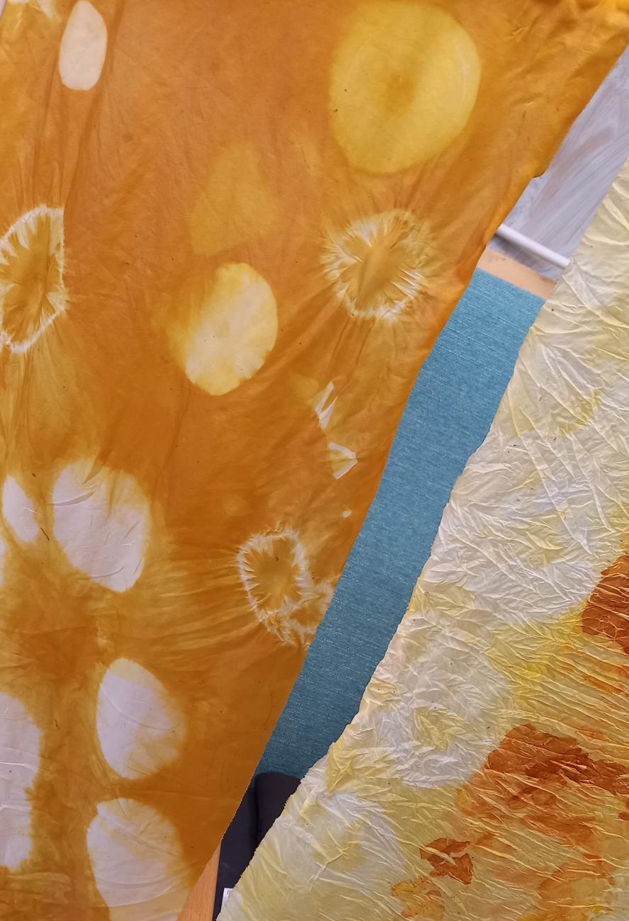 Two strips of fabric are hanging up together. On one, tie dyed spots sit softly on the yellow orchre background, like jellyfish floating. The other fabric has golden and orange patches and has an interesting crinkled texture.