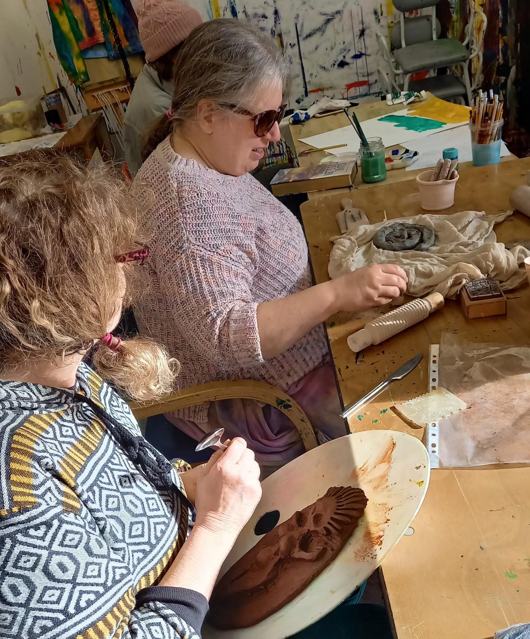 Two people are sitting down at a big table making things with clay. They are both busy an using their hands to mold the clay into shapes.