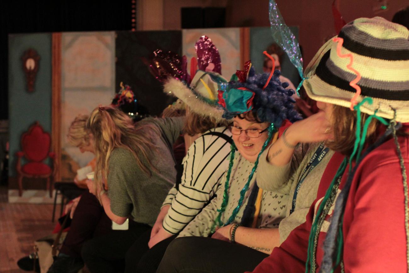 People are sitting in a row watching a fashion show. They are wearing colourful hats and bright clothes and smiling. The background is dark.