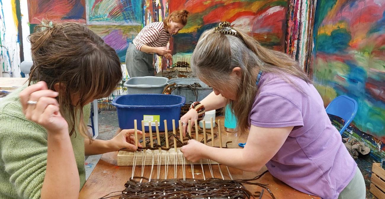 Two people are winding strips of seaweed around poles in a peg loom. They look busy and interested.