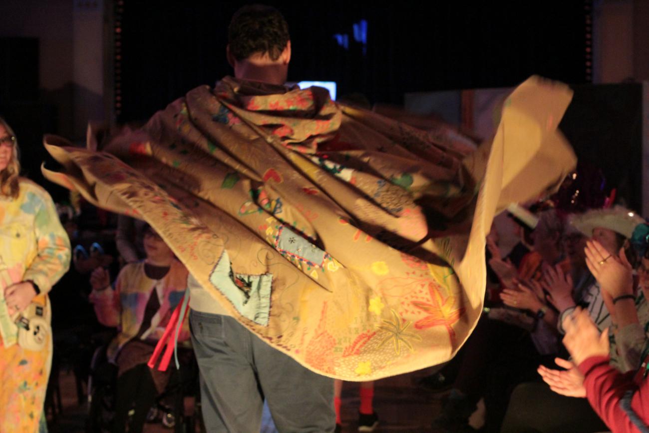 A big brown cape is swirling before the camera as a person turns on the catwalk. The cape is adorned with colourful patterns, images, ribbons and feathers. At the sides we can see the hands of people clapping in the audience.