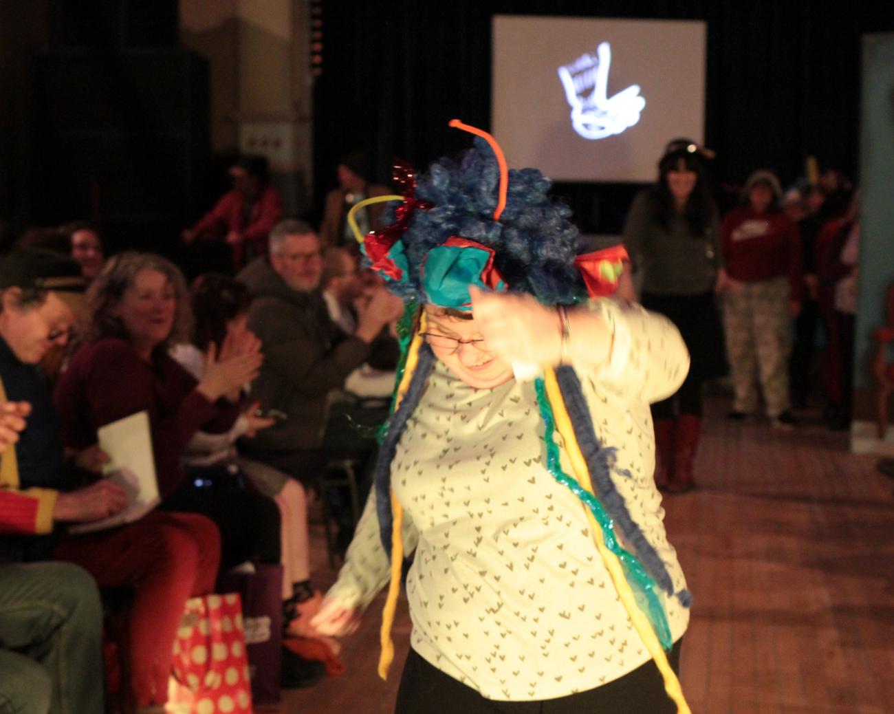 A person is dancing in the foreground wearing a white top with little grey hearts on it and a blue wig adorned with yellow, turquoise and red fabric and tissue paper. She has one arm up and is looking down and smiling.