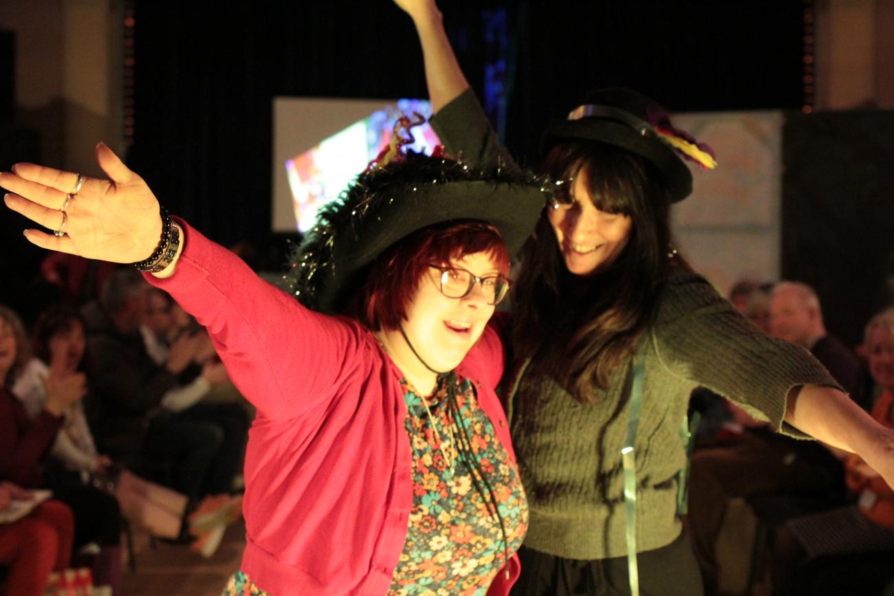 Two people are standing in front of the camera together. The one on the left is wearing a pink cardigan and a pink and green flowery top and the one on the right is wearing a dark green sweater. They are both holding their arms out, smiling and wearing black sparkley hats. Behind them there is an audience and a projected white image.