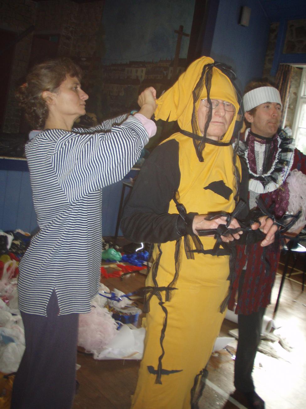 3 people standing in a hall space. One person is wearing a yellow costume that is long and thin with black ribbons coming of it. Another person is tying ribbons onto the person in yellow. The 3rd person is wearing a purple costume with a white tennis like headband. 