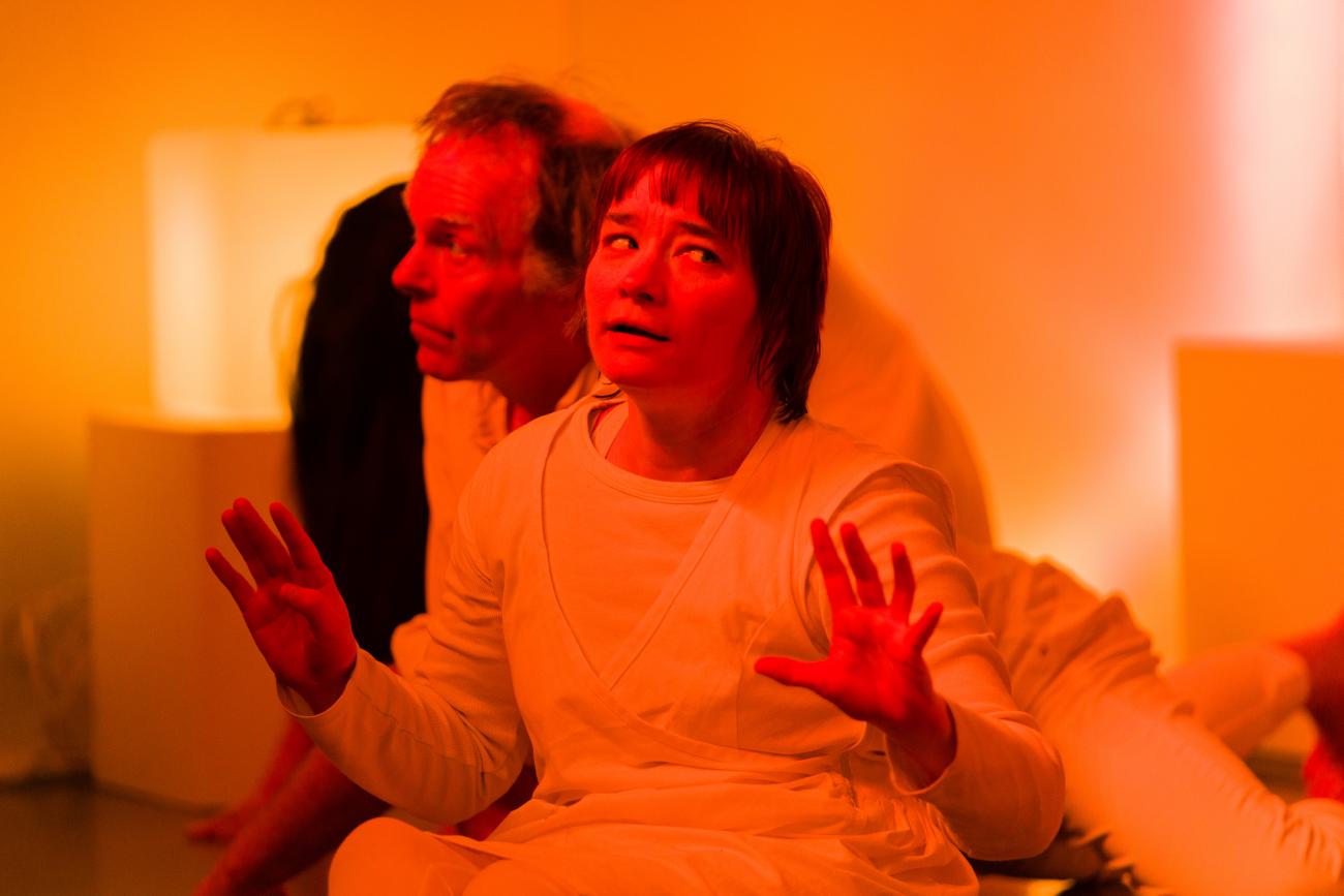 2 people dancing together in an orange lit space. They are both wearing white. One person has their hands out and open and is looking to the right. The other person is sitting behind them and is looking to the left