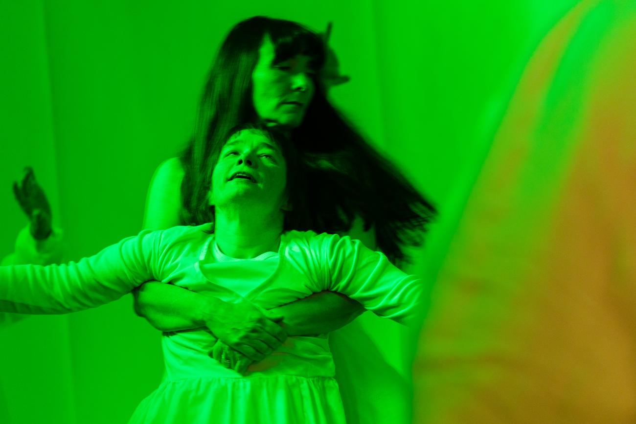 2 people dancing in green light. They are both wearing white. One is holding the other from behind through their arms. There person being held has their arms open and is looking upwards.