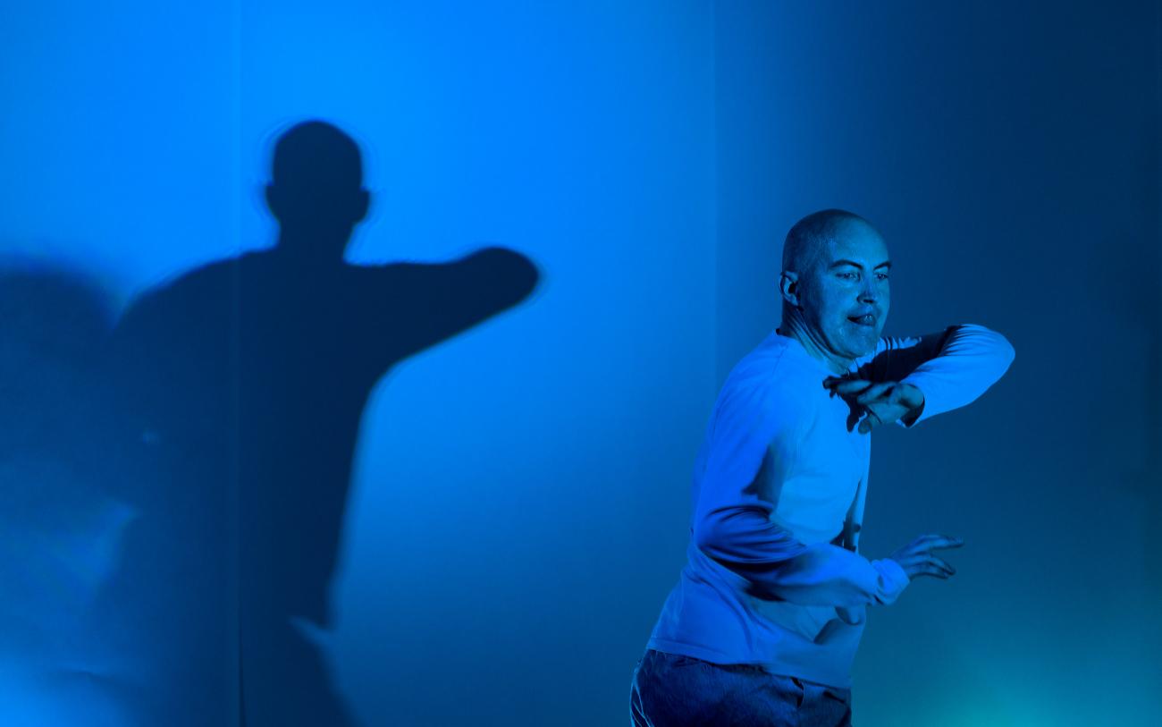 a person dancing in a blue lit space. They are wearing white and they are moving their elbows outwards. They are casting a huge shadow of their silhouette on the wall behind them