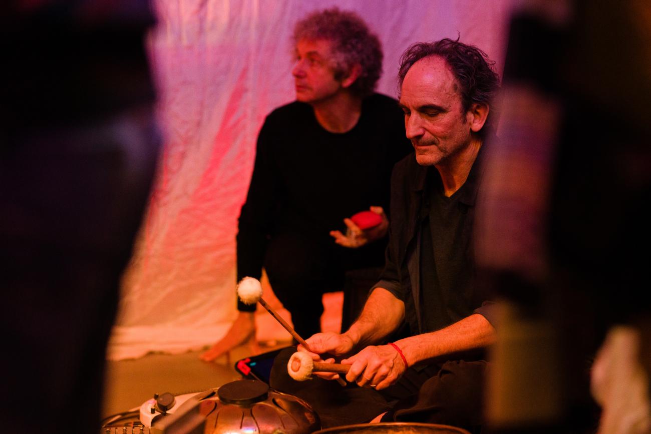 A person sitting on the floor in a busy space playing percussion instruments.