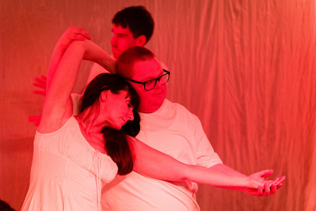 3 people dancing in a red space. All of them are wearing white. 2 people are dancing close together with their arms around each other. The third is in the background.