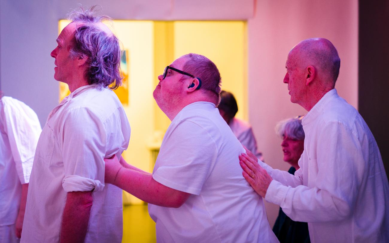 3 people dressed in white performing. They have their hands on each others backs and are standing in a line
