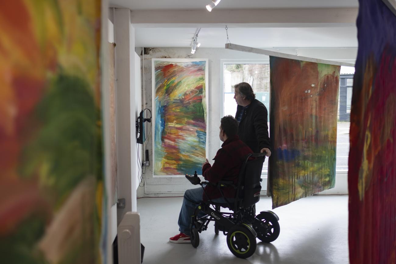 colourful abstract paintings hanging in gallery with 2 people looking at the paintings. One person is in a wheelchair and one person is standing