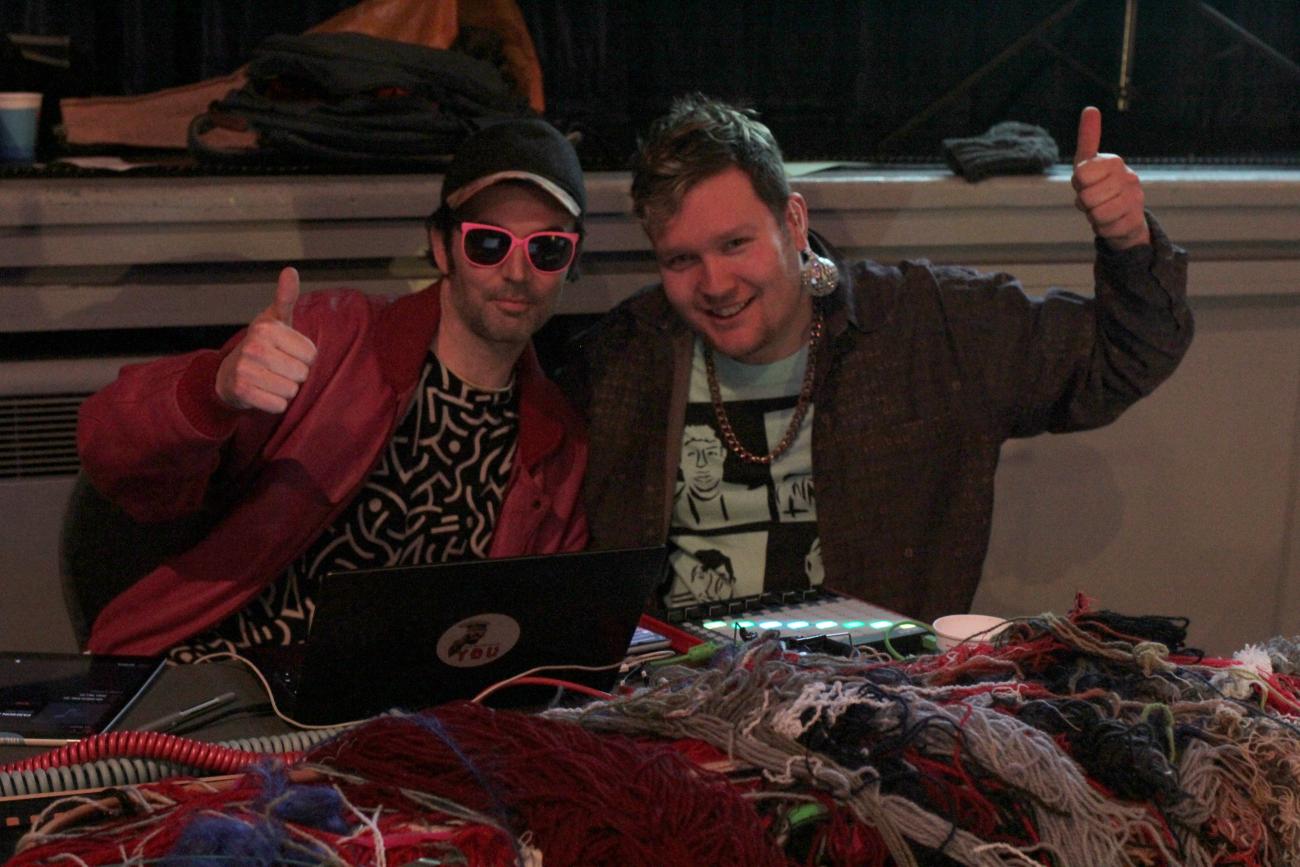 Two people are posing for the camera with an arm around each other with the other arm doing a thumbs up. They are sitting in front of a laptop and a sound mixer. One is wearing pink sunglasses and the other is wearing mirror ball earrings.